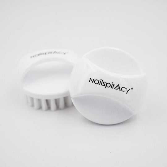 Two white, round-shaped nail dust brushes with bristles, one standing upright and the other lying on its side, with the brand name 'Nailspiracy' visible on the side of the standing brush.