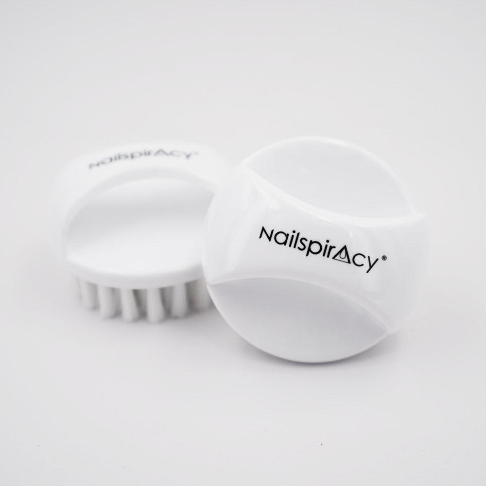 Two white, round-shaped nail dust brushes with bristles, one standing upright and the other lying on its side, with the brand name 'Nailspiracy' visible on the side of the standing brush.